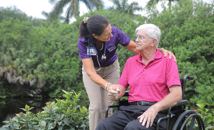 A caregiver holds the hands of a woman who is sitting up in bed, while both talk with a VITAS physician